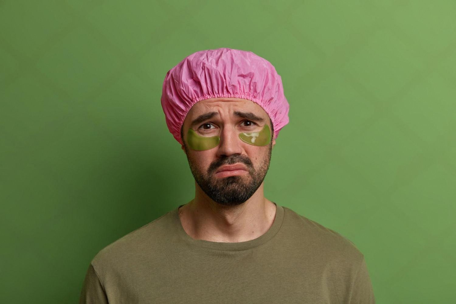 Tired man with pink shower cap and green under-eye patches showing dark circles